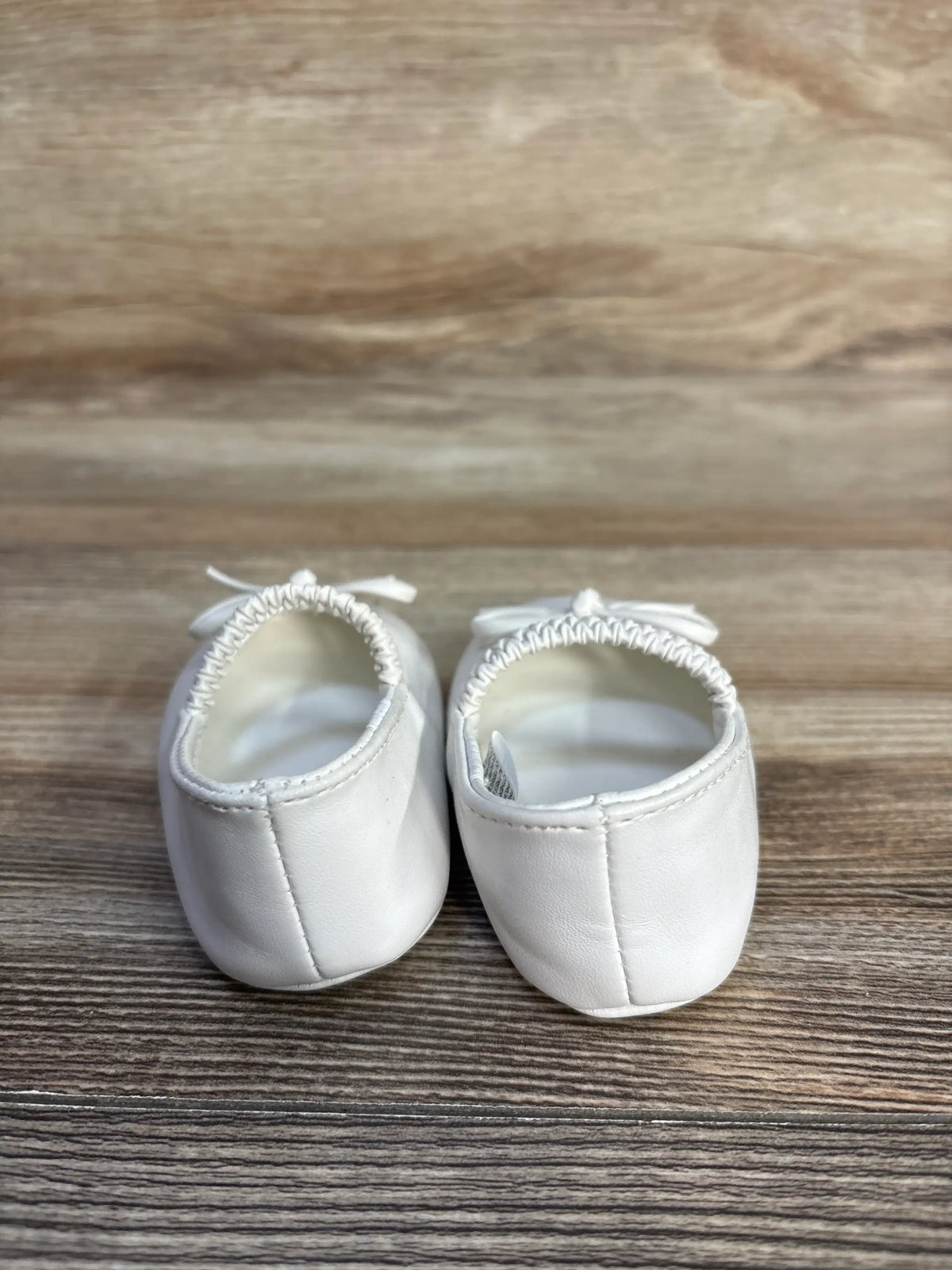 Pair of white Janie & Jack bow soft sole ballet flats for toddlers on a wood surface.