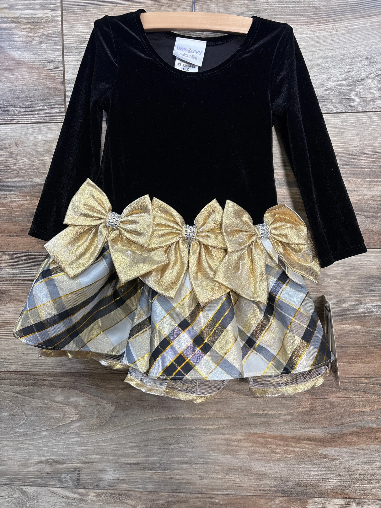 Front view of Iris & Ivy black velvet toddler dress showing long sleeves, three gold bows and gold-gray plaid skirt on wood floor