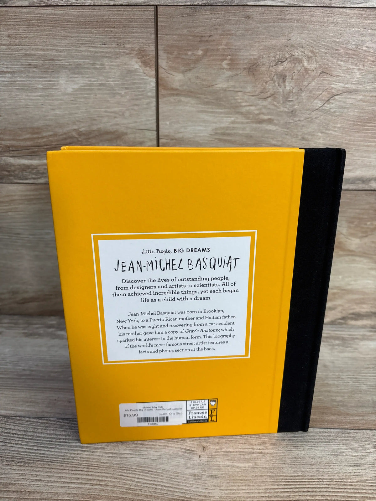 Back cover of Jean-Michel Basquiat hardcover book with Little People, BIG DREAMS text, yellow cover, black spine, and wood surface