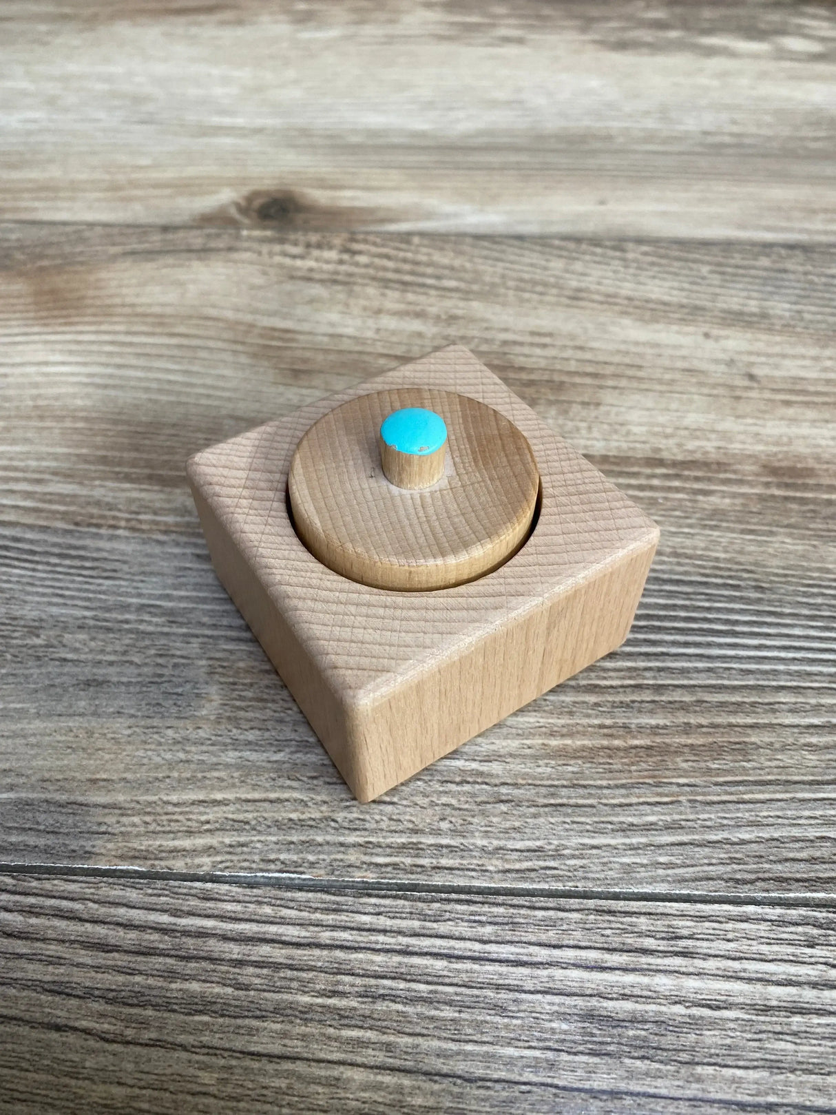 Angled view of Lovevery Pincer Puzzle showing a natural wood square base and removable round knob with turquoise dot