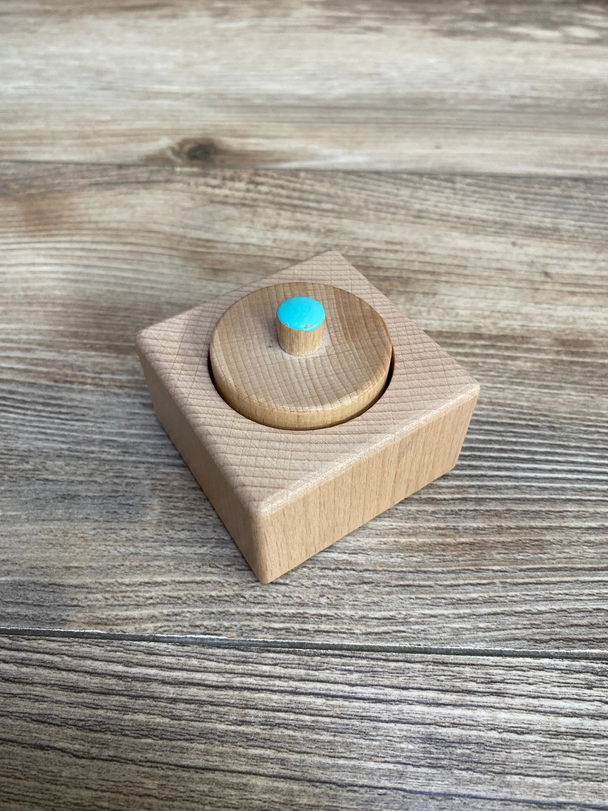 Angled view of Lovevery Pincer Puzzle showing a natural wood square base and removable round knob with turquoise dot