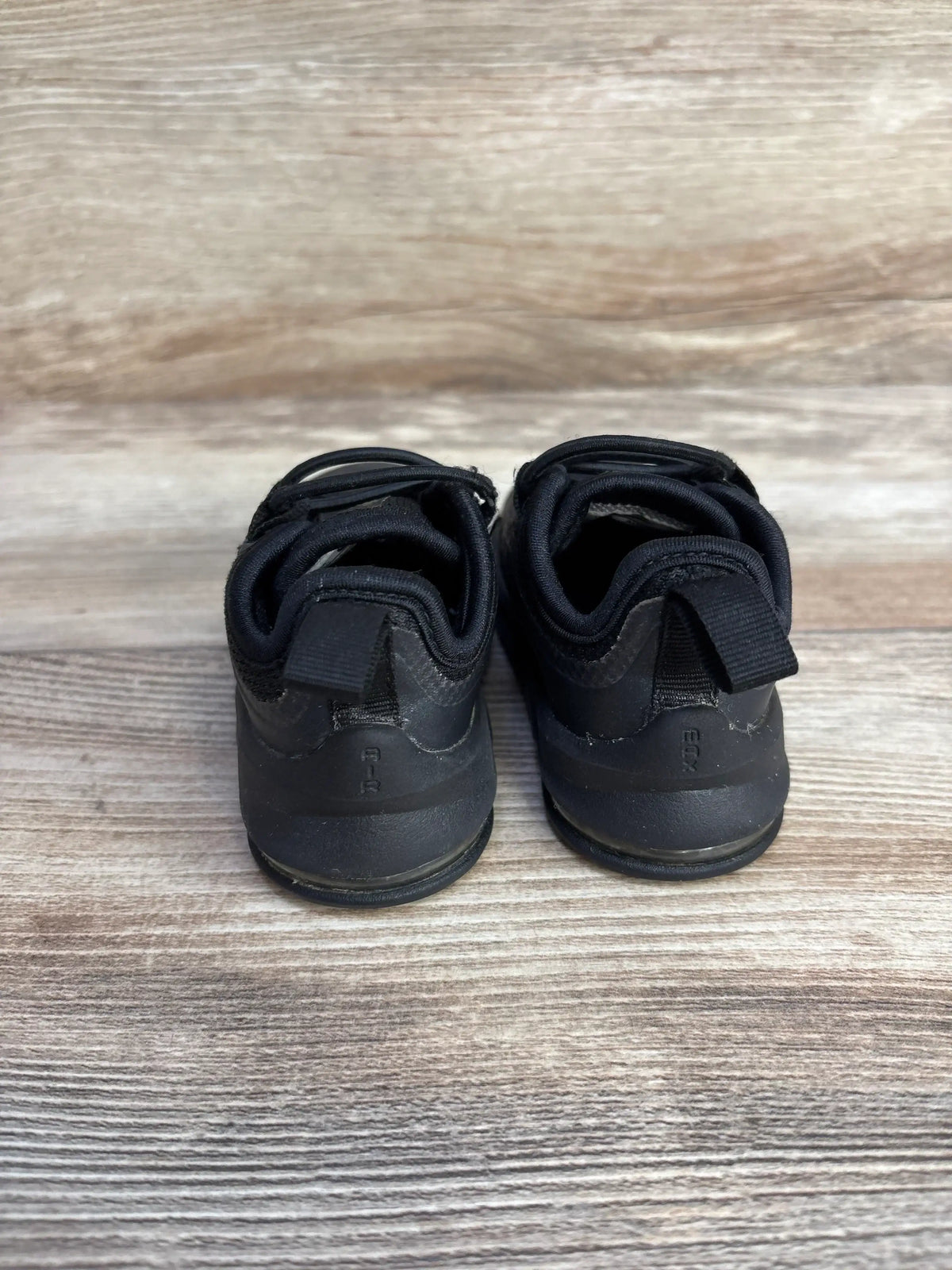 Rear view of toddler black Nike Air Max Axis sneakers showing heel pull tabs and light scuffing on wood plank background