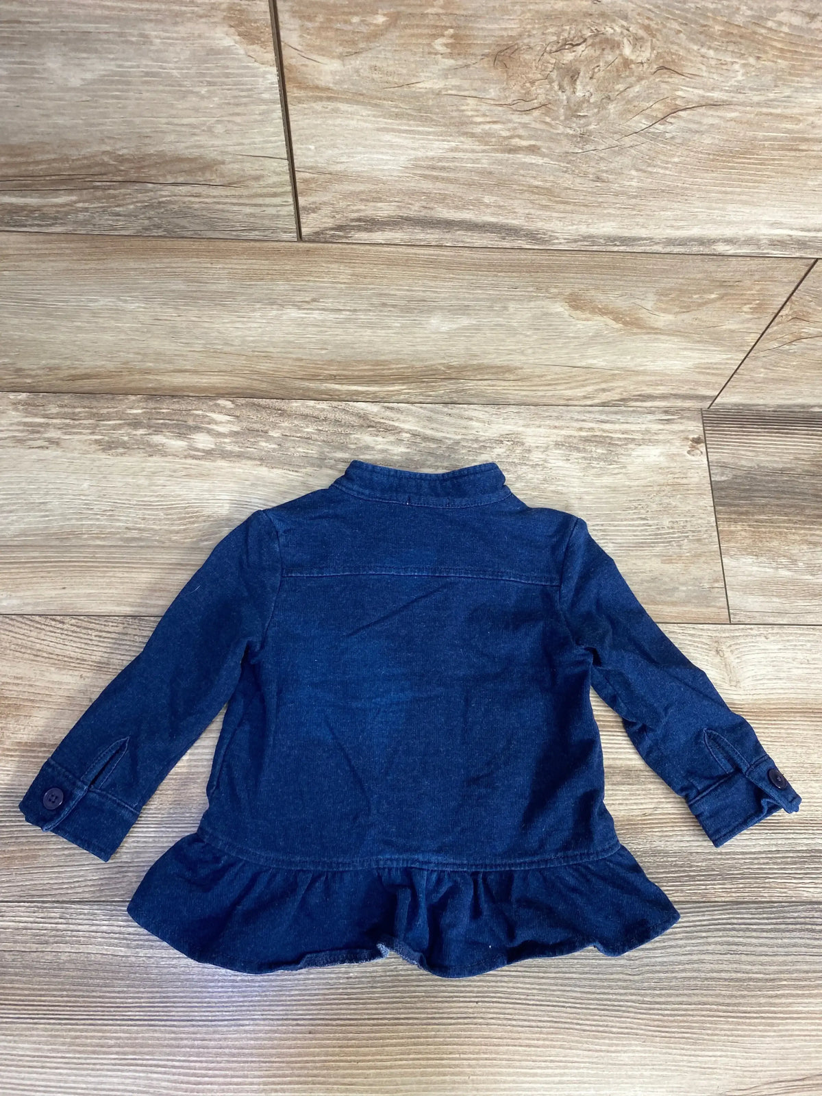 Back view of a navy blue denim jacket with ruffle hem and buttoned cuffs on wooden floor
