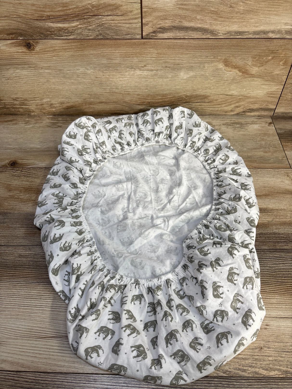 Top-down view of Bublo Baby fitted bassinet sheet showing gray elephant print and elastic gathered edges on wood floor background