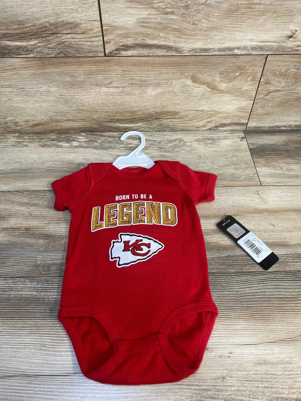Red baby bodysuit with Born to Be a Legend and football logo on wooden floor.