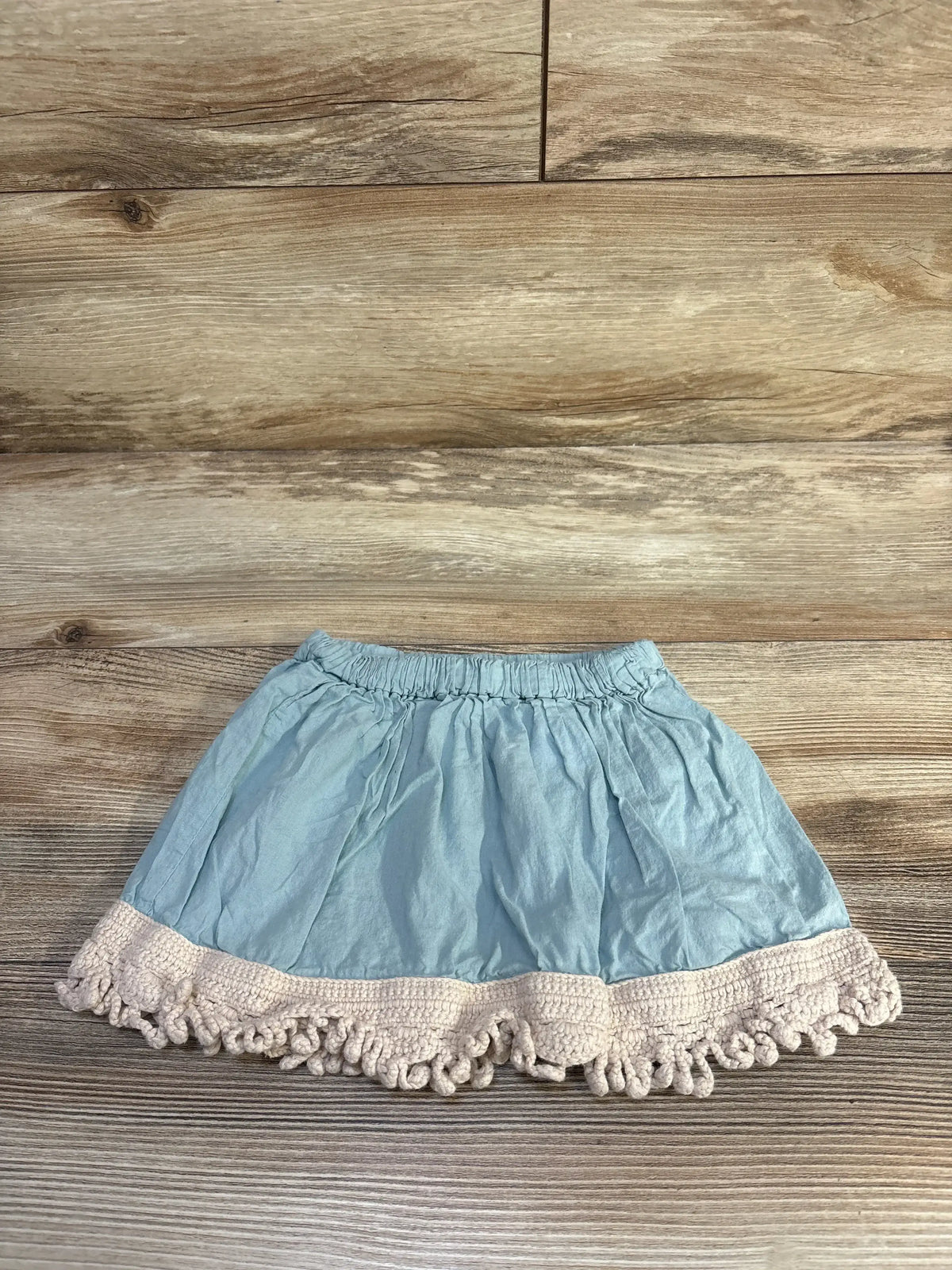 Flat lay of blue Kate Quinn toddler skirt showing elastic waistband and cream crochet scalloped hem on wood floor background