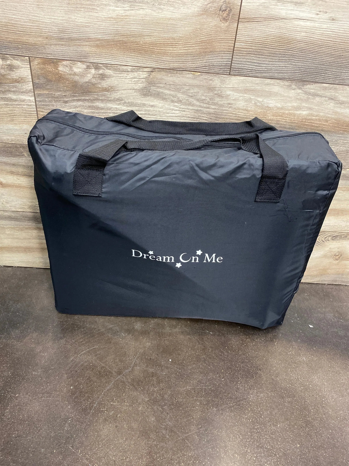 Folded side view of Dream On Me Travel Light playard in black in zippered carry bag showing handles against wood wall