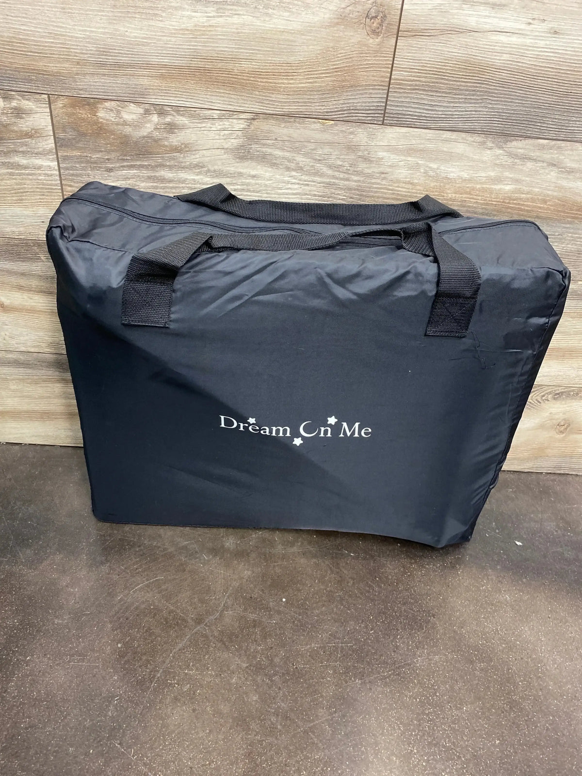 Folded side view of Dream On Me Travel Light playard in black in zippered carry bag showing handles against wood wall