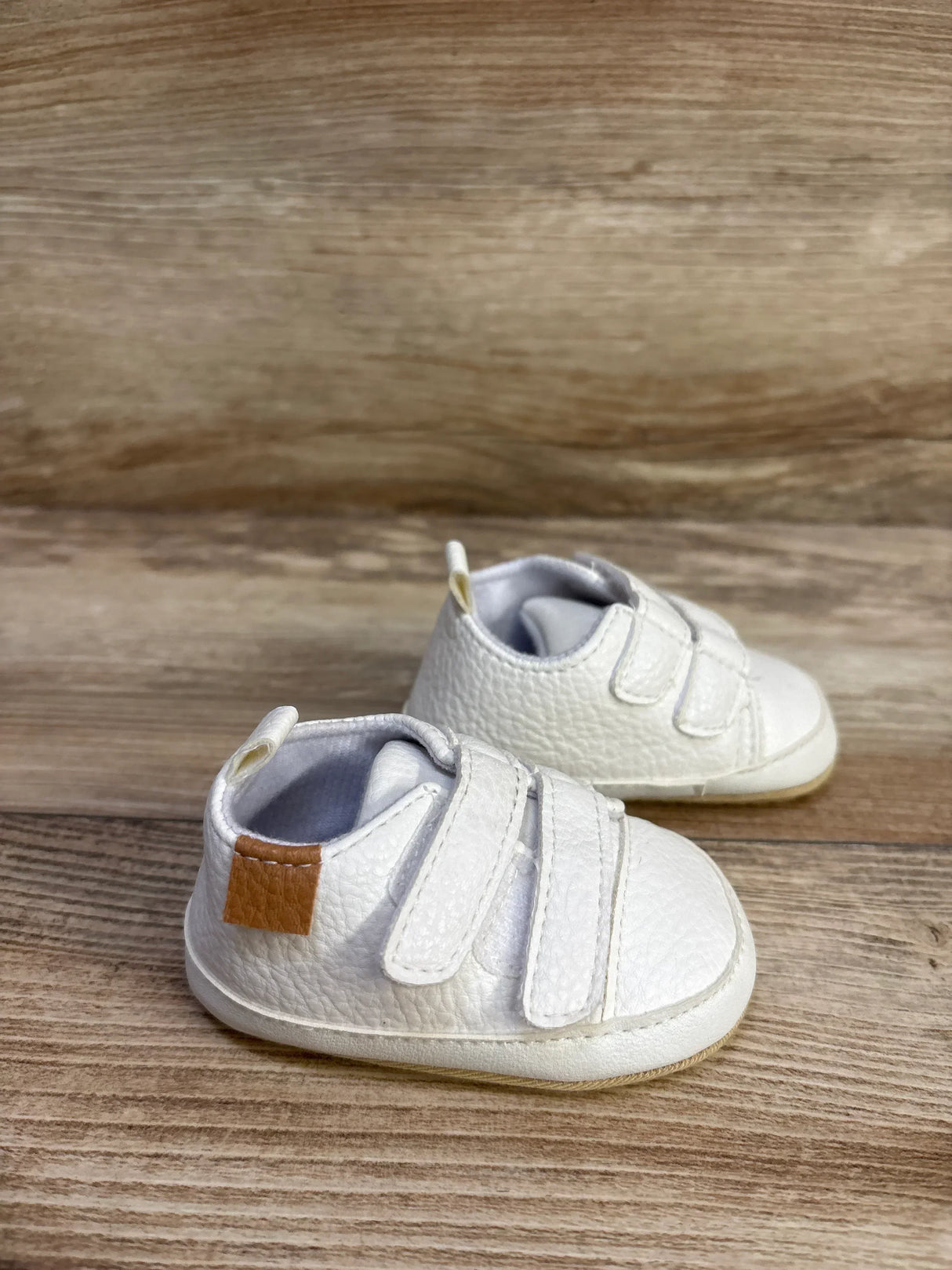 Three-quarter view of Myggpp baby first-walker sneakers in white showcasing two Velcro straps, textured leather-look upper and tan heel patch on wood background