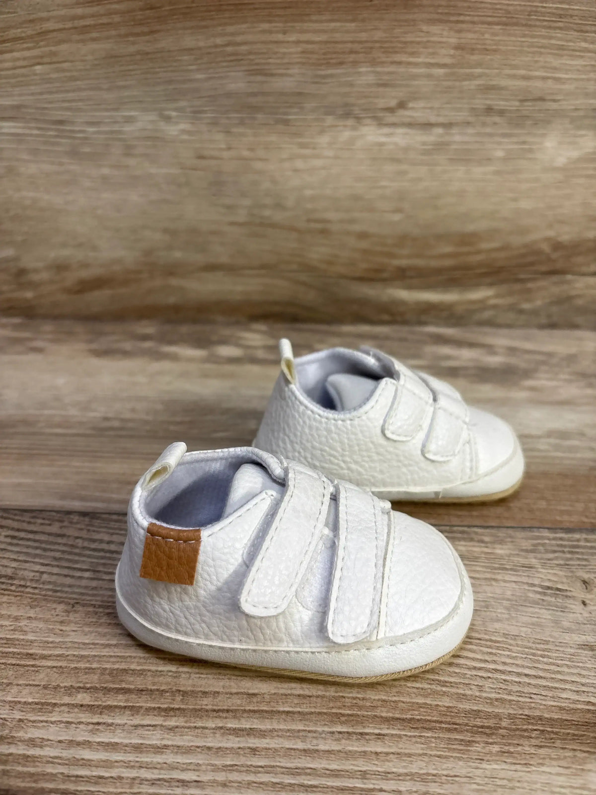 Three-quarter view of Myggpp baby first-walker sneakers in white showcasing two Velcro straps, textured leather-look upper and tan heel patch on wood background