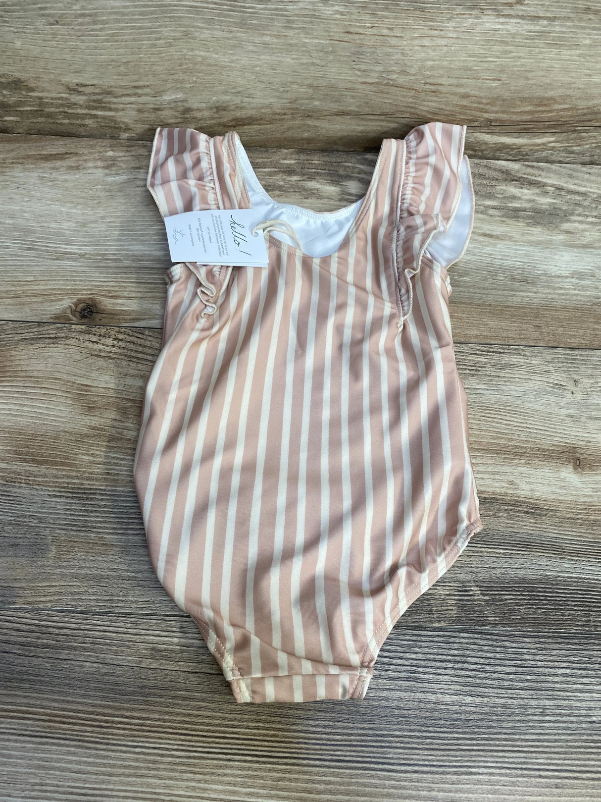 Flat-lay of Pehr ruffle swimsuit showing pink and white vertical stripes, ruffled cap sleeves, and attached tag on wood background