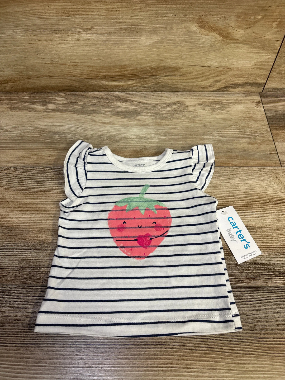 Flat lay Carter’s baby striped white and navy shirt with smiling strawberry print and ruffle sleeves on wood floor, size 3M tag