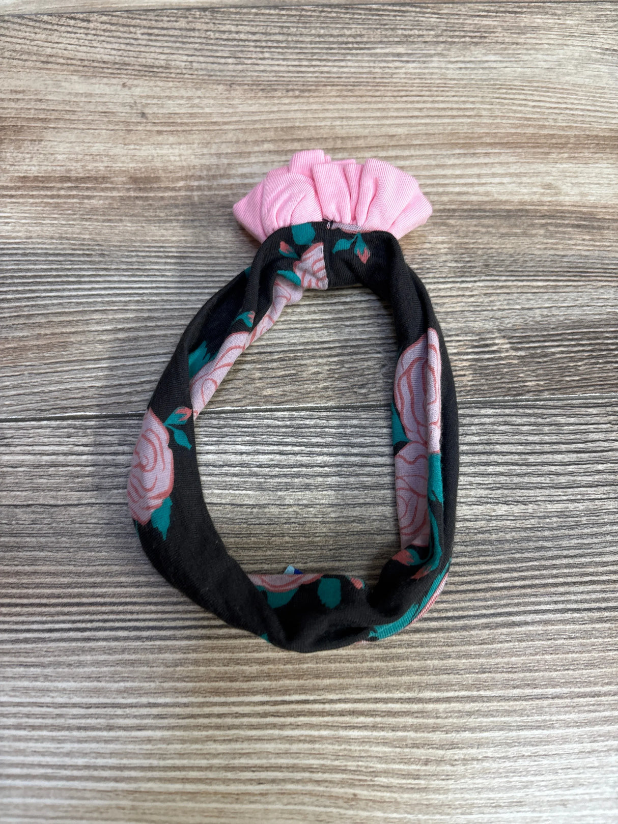 Top-down view of Kickee Pants baby headband showing black fabric with pink rose print and pink ruched knot on wood background