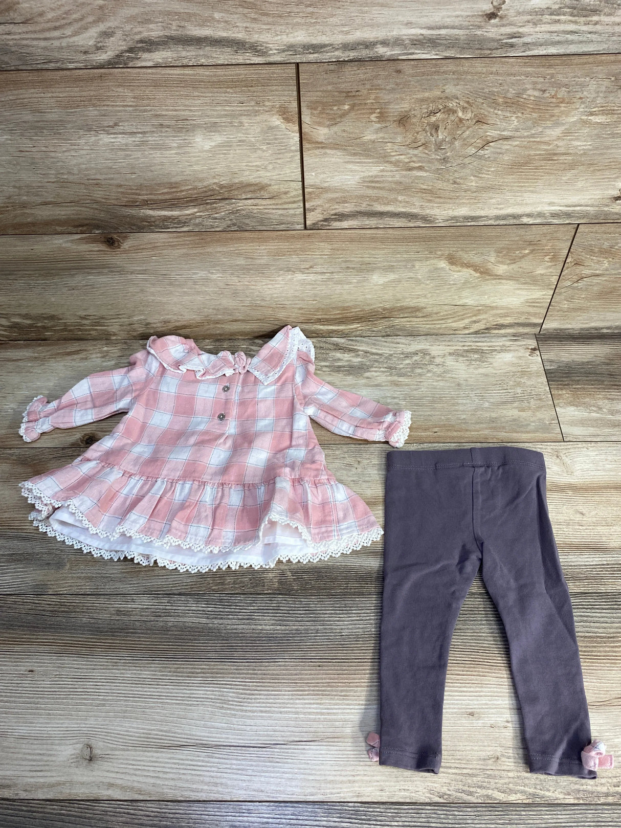 Flat lay of baby girl 2-piece pink plaid top and gray leggings showing lace trim, ruffled collar, and ankle bows on wood background