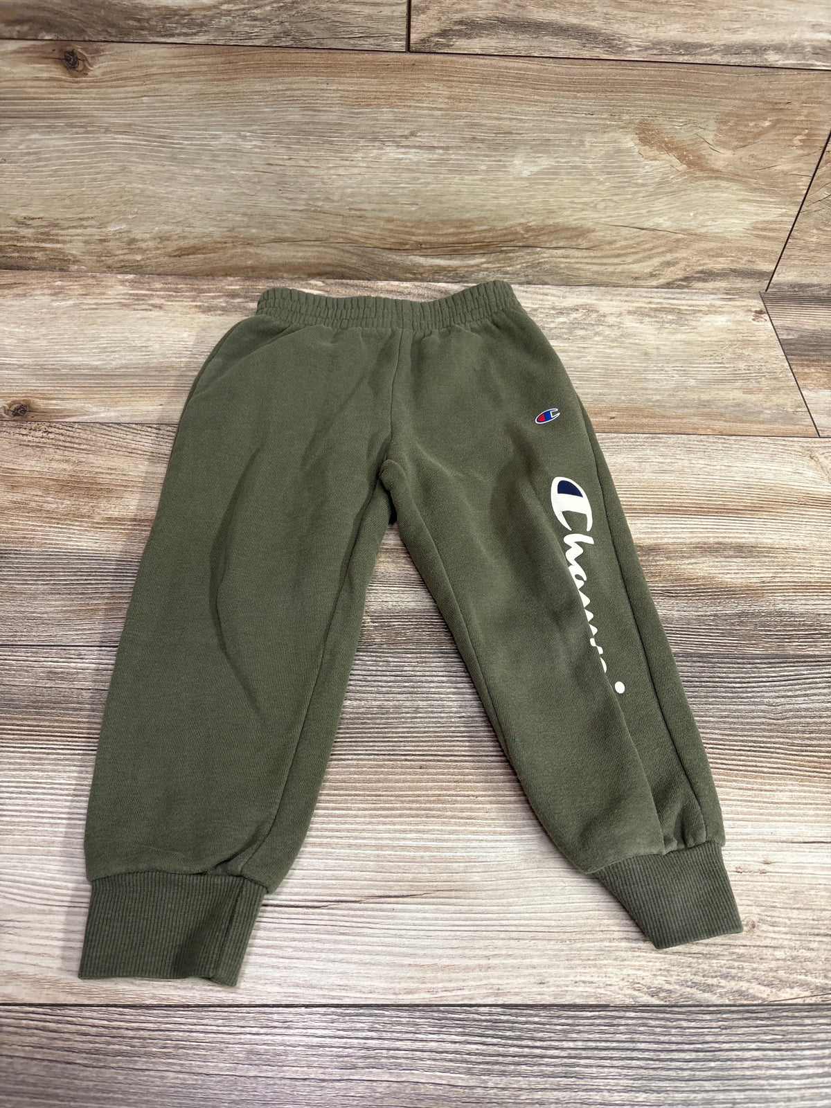 Top-down view of green Champion joggers for size 3T showcasing cuffed ankles and logo on a wood-grain background