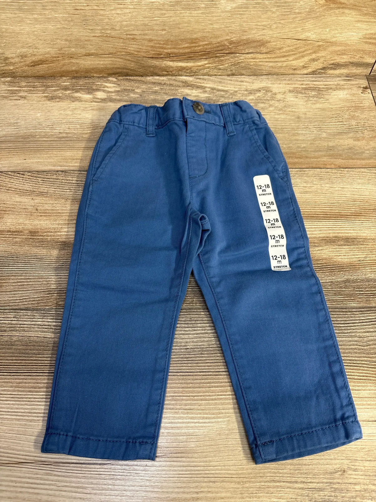 Flat-lay Children’s Place blue toddler pants with elastic waist button, pockets and size sticker ‘12-18 m’ on wood