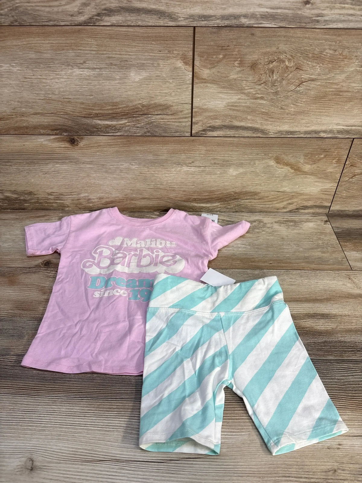 Flat lay of toddler two-piece: pink shirt with Malibu Barbie graphic and mint-and-white striped shorts on wood floor