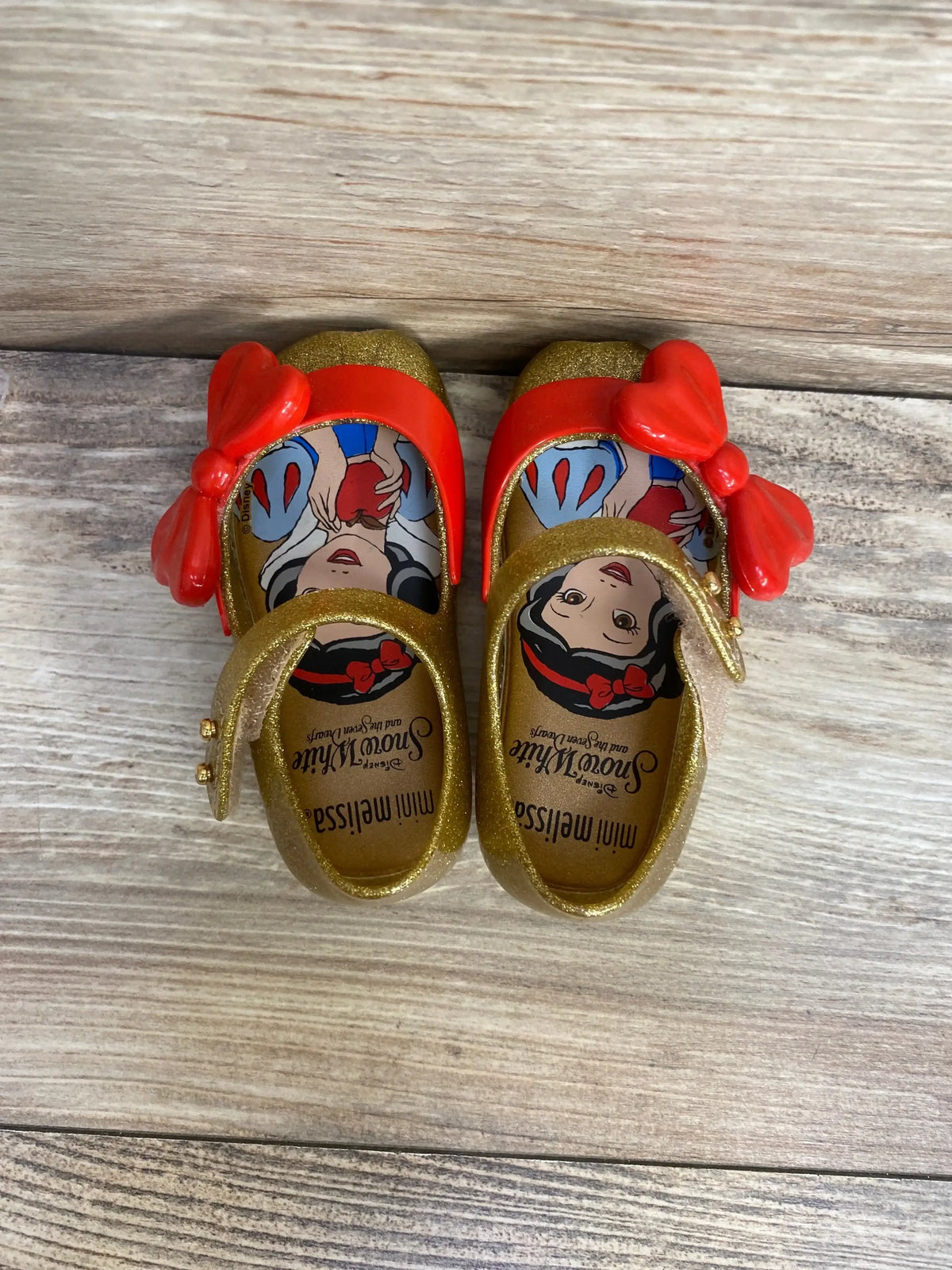 Top view of Mini Melissa Ultragirl Snow White toddler shoes showing gold glitter finish, red bows and Snow White insoles on wood background