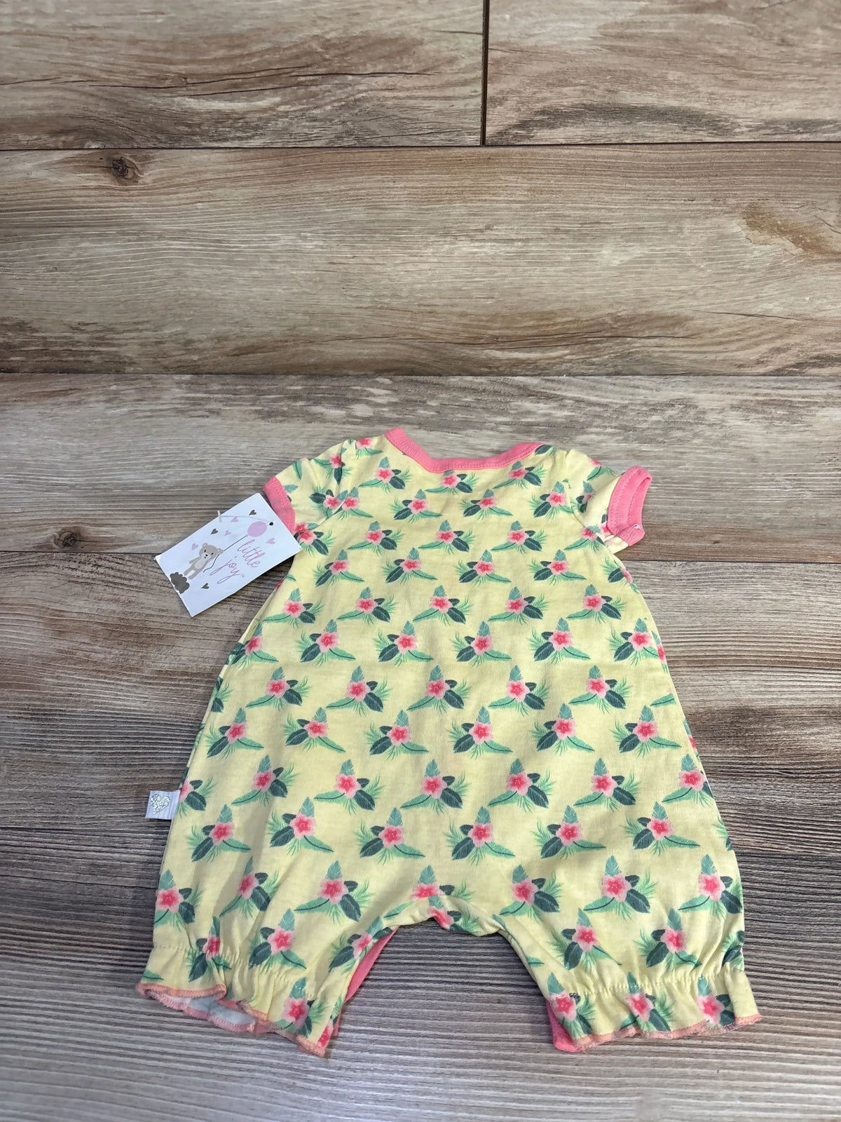 Flat lay of yellow shortie romper with pink floral print, pink trim, elastic leg openings, and brand tag on wood background