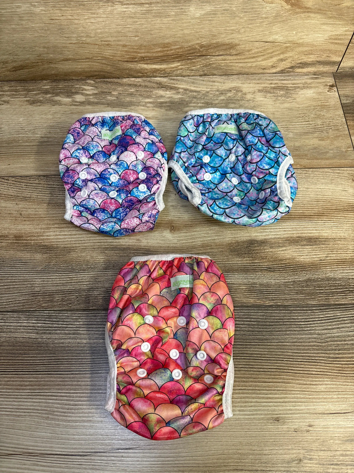 Top view of three Wegreeco reusable swim diapers in colorful prints with adjustable snap closures on a wood background