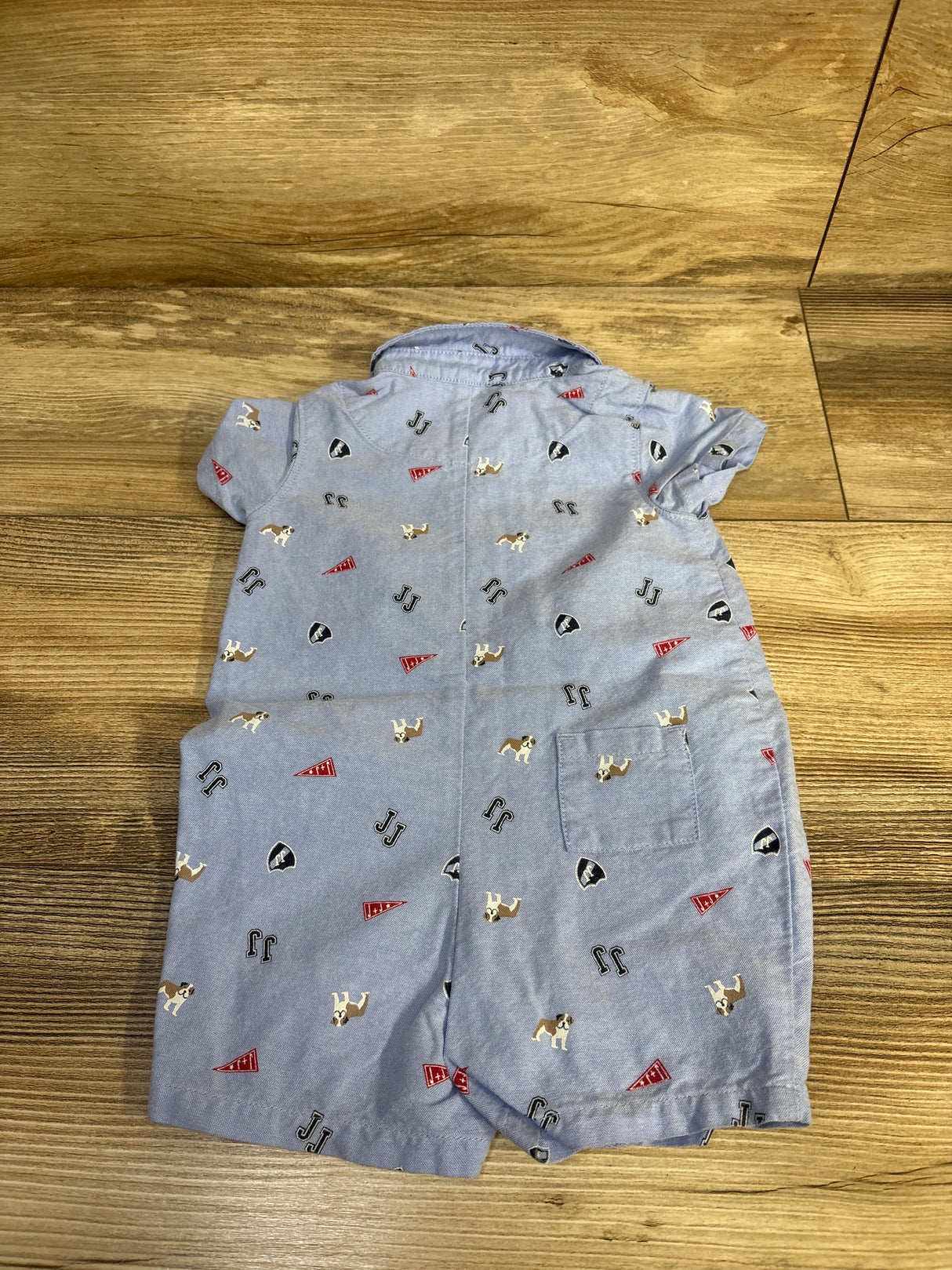 Flat-lay back view of blue oxford baby romper with bulldog and pennant print, short sleeves and back pocket on wood background
