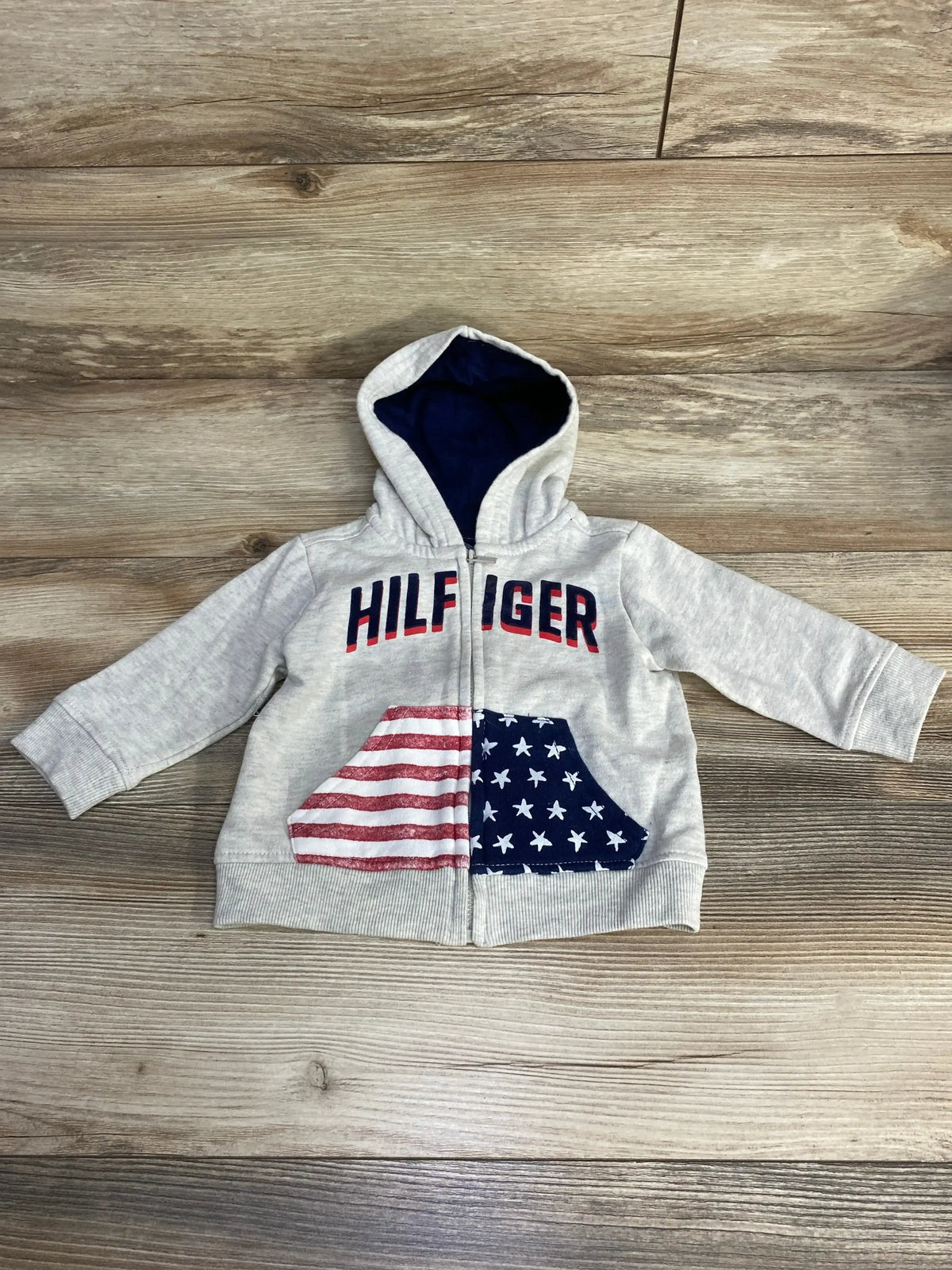 Flat-lay of grey Tommy Hilfiger baby zip hoodie with navy hood lining, HILFIGER lettering, and American-flag pouch on wood