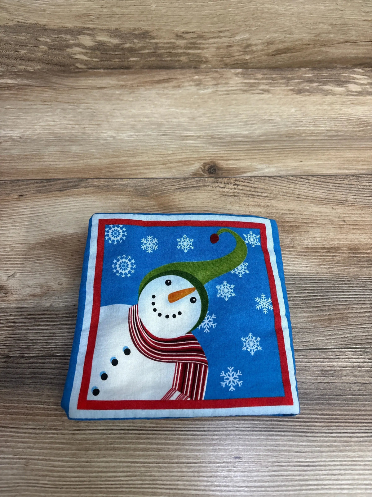 Top-down of A Snowman Story soft cloth book showing snowman with green hat and red striped scarf on wood surface