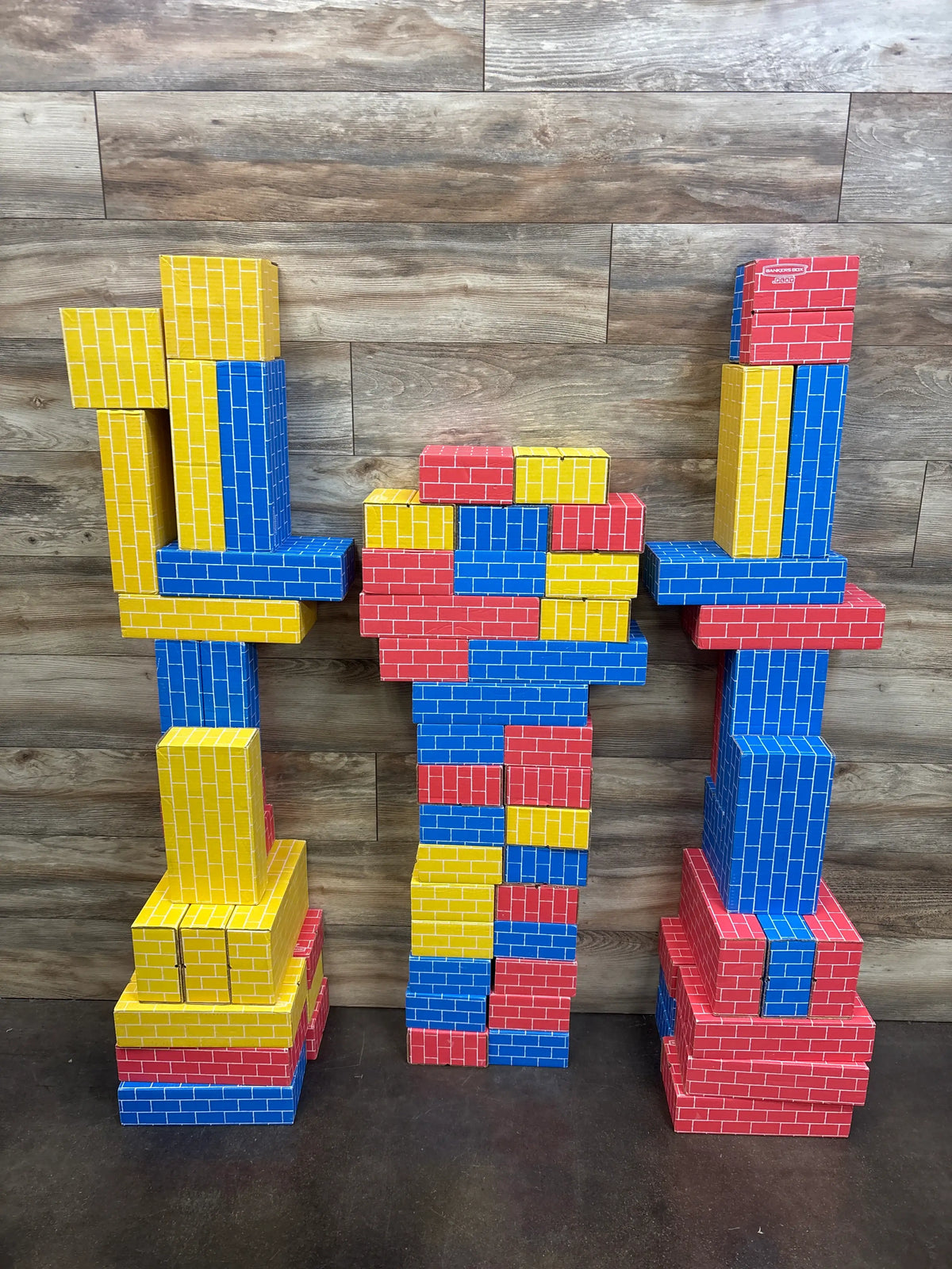 Front view of Bankers Box Heavy Duty Jumbo Cardboard Building Blocks 74ct arranged as red, blue, and yellow towers on dark wood