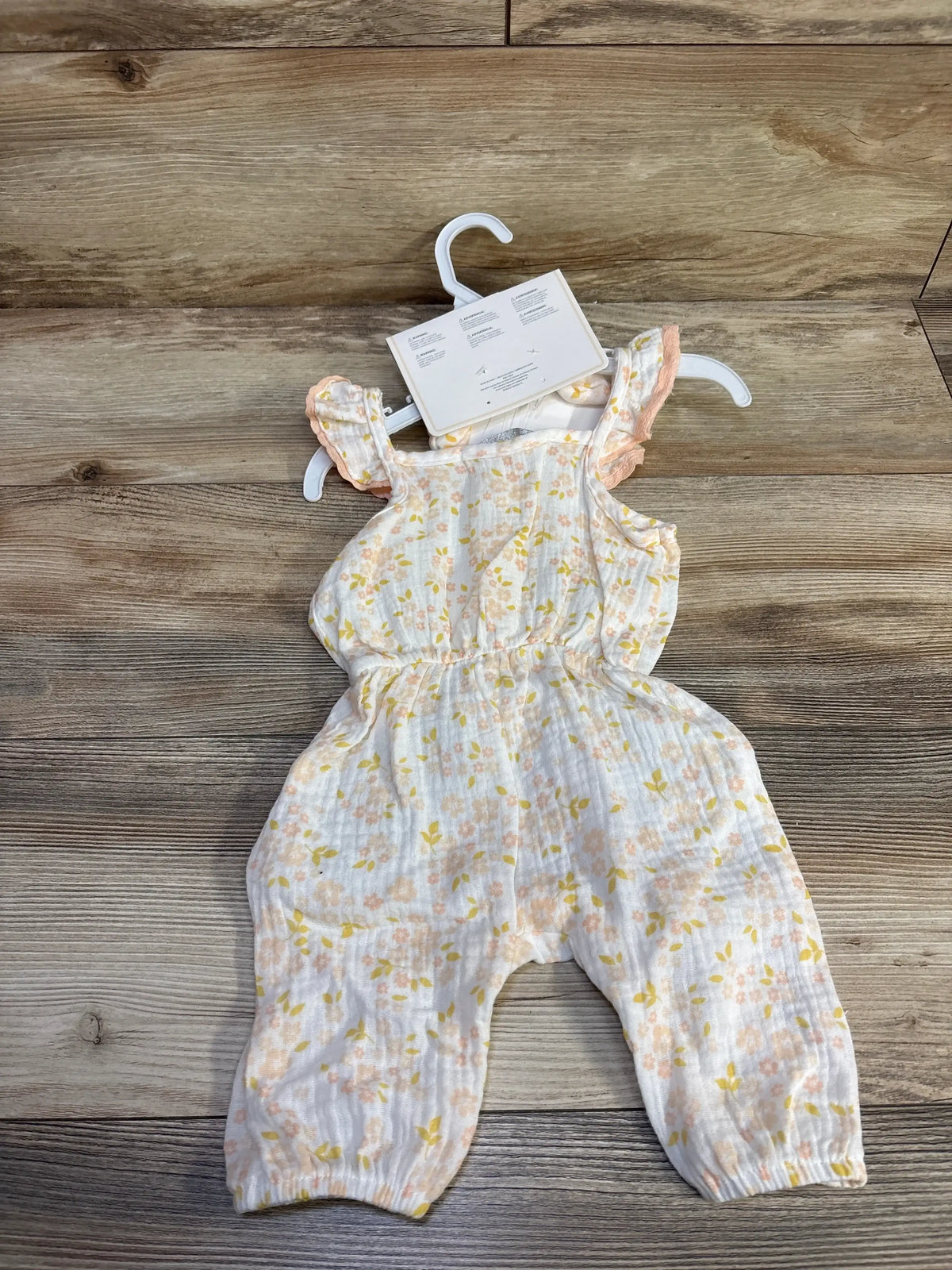 Top-down view of newborn white gauze jumpsuit showcasing ruffled shoulder straps and peach and yellow floral print on wood background