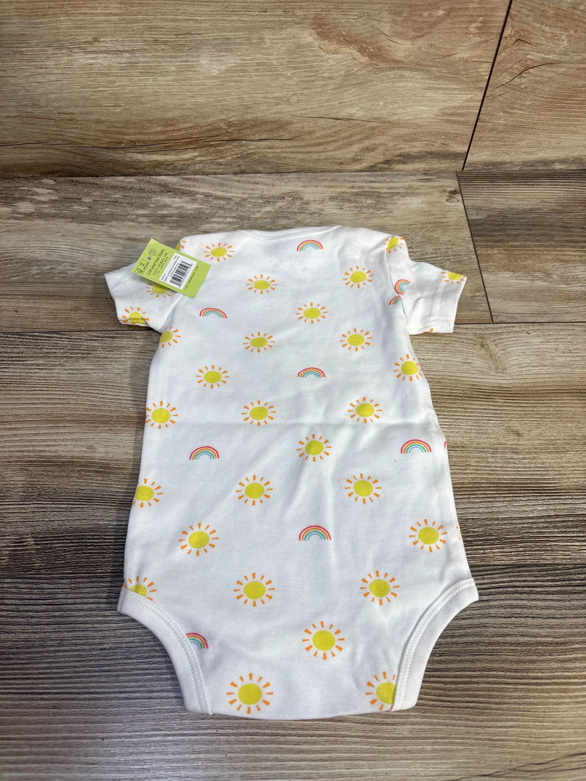 Front view of a white Primary baby bodysuit in size 3-6 months showcasing sun and rainbow print on a wood-look background