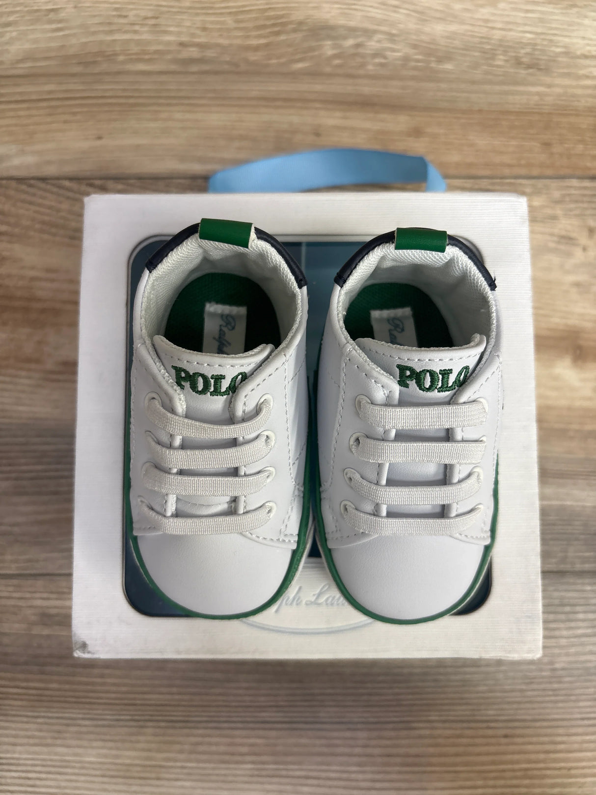 Pair of white and green Polo baby shoes with laces, displayed on a white box on wooden surface