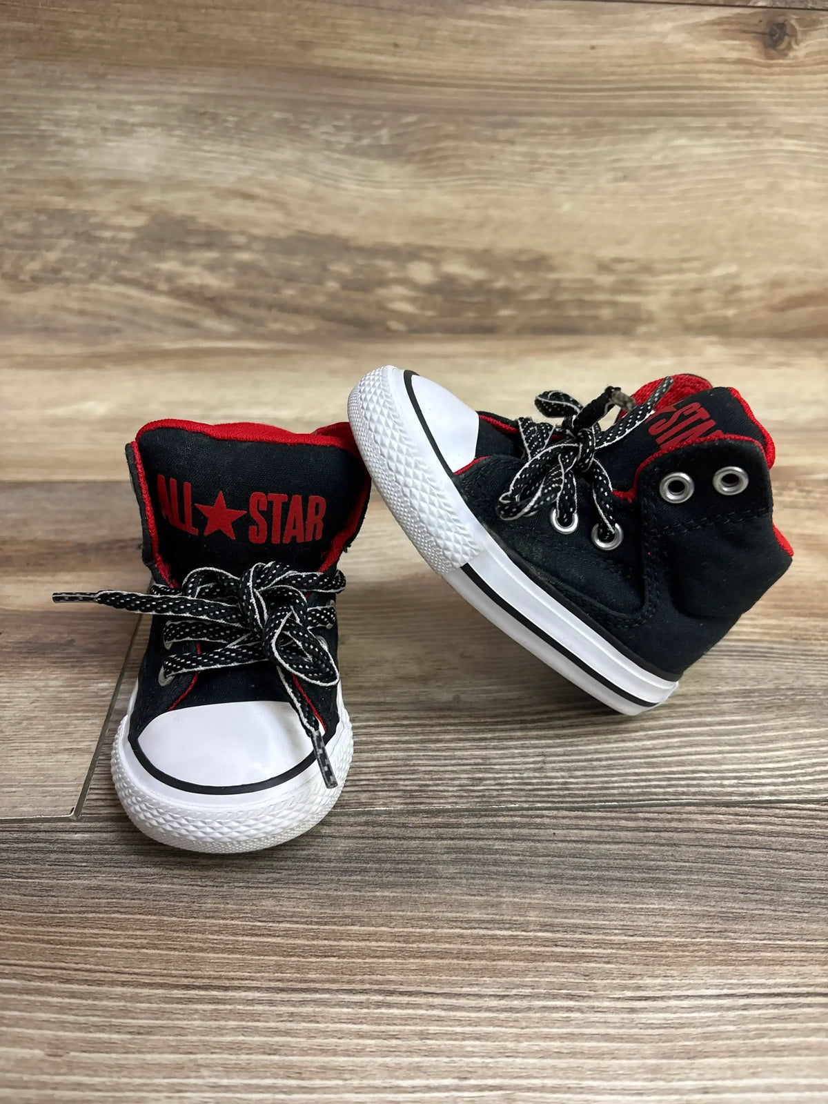 Three-quarter view of All Star toddler high-tops in black canvas with red logo panel and white rubber soles on wood floor
