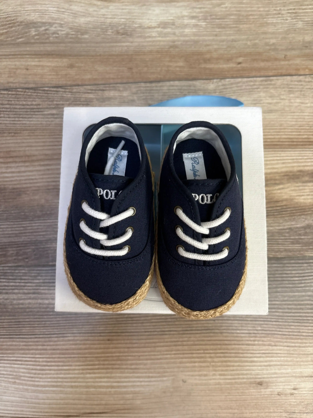Top view of Ralph Lauren Bowman lace crib shoes showcasing navy fabric, white laces, and tan rope soles on a white box