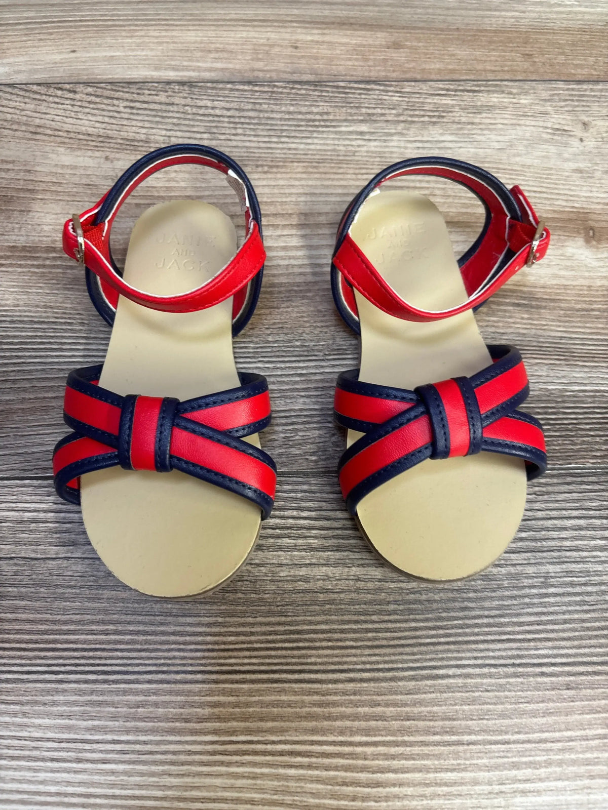 Top-down view of toddler Janie and Jack sandals in very cherry red and navy, crossed double straps and ankle buckle on woodgrain background