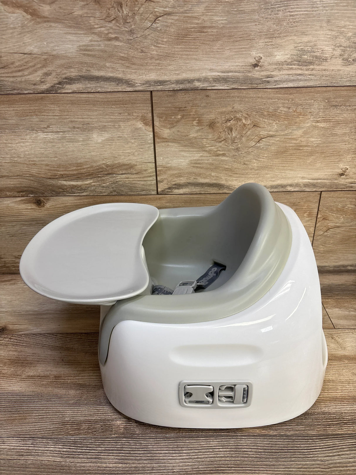 Three-quarter view of Bumbo Multi Seat in grey with removable tray attached and harness buckles visible on a wood plank background