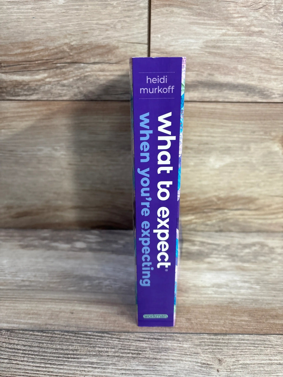 Spine view of What to Expect When You’re Expecting 5th Edition paperback book showing purple binding on wood-grain background