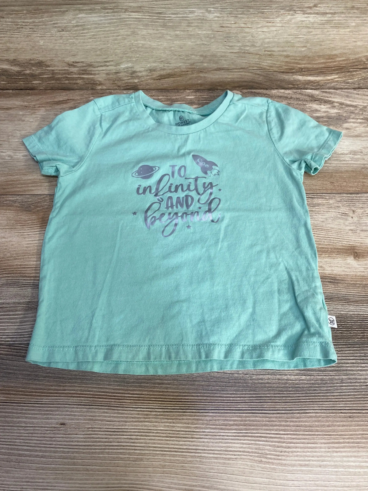 Flat lay of mint green toddler short-sleeve shirt (4T) showing "To infinity and beyond" script with rocket and planet graphics on wood background