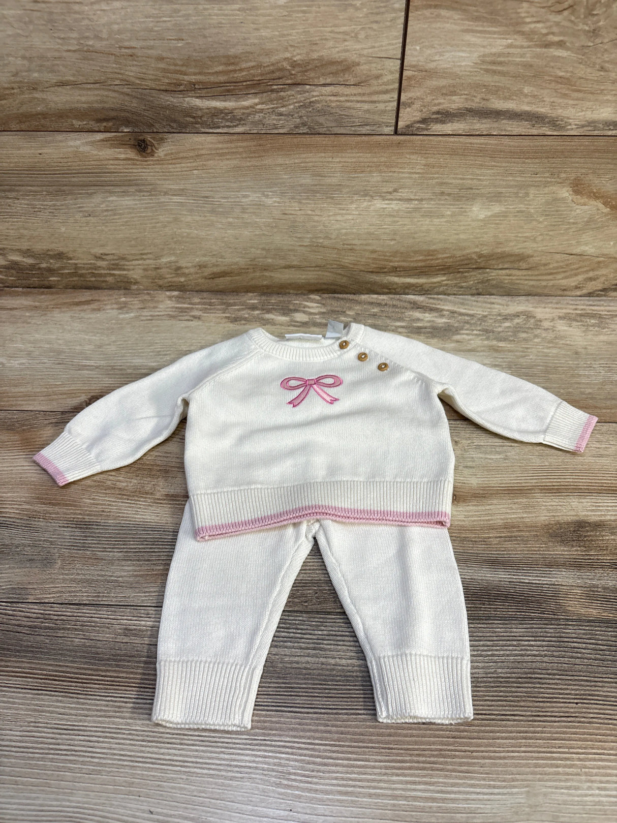 Flat lay of ivory knit 2-piece sweater and pants with pink bow and trim, three shoulder buttons on wood background