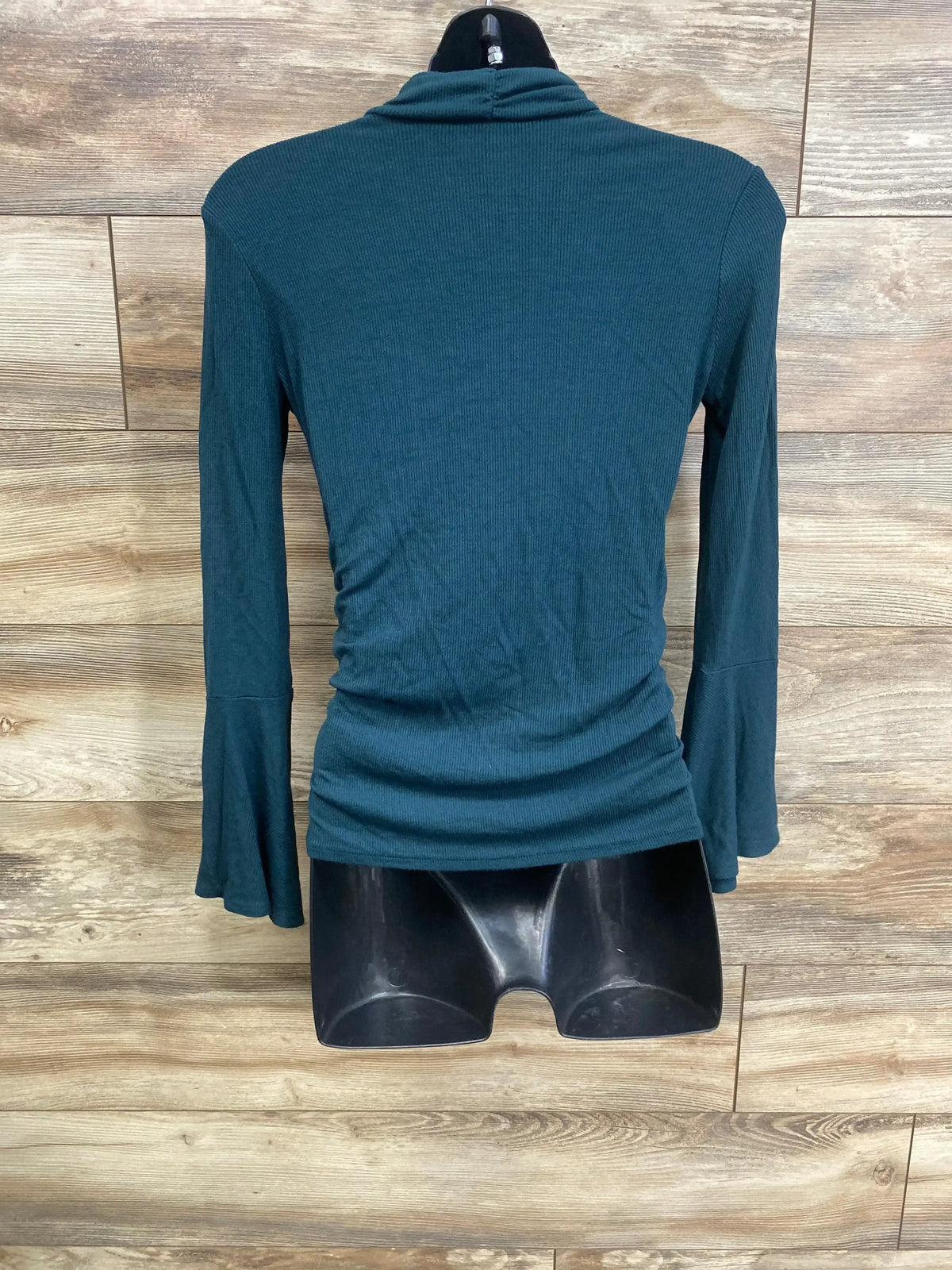 Back view of maternity ribbed ruched turtleneck shirt in green showing side ruching and flared sleeves on wood backdrop