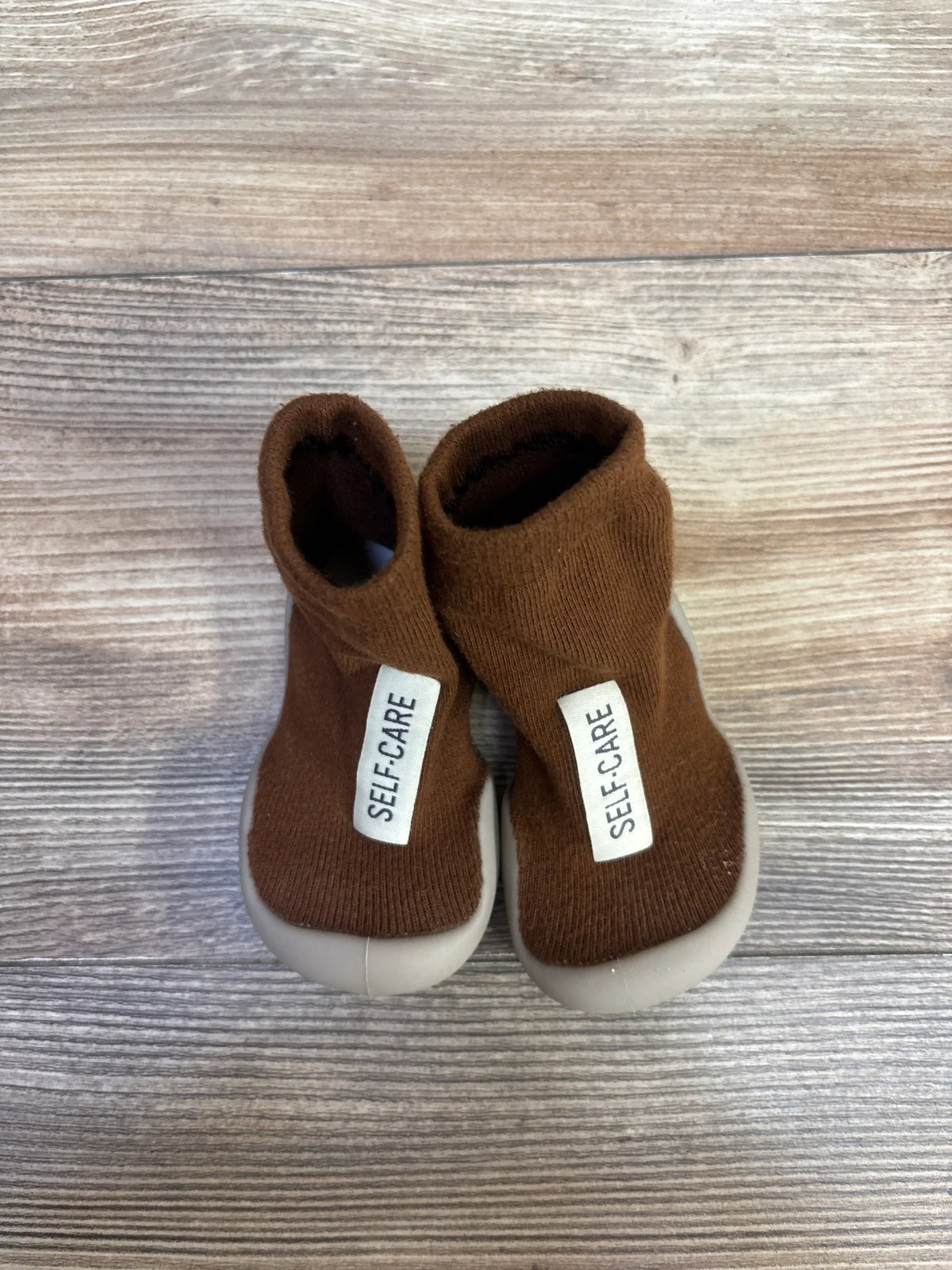 Front view of Tiny Explorings Self-Care sock shoes in brown, size 2/3C, with cream soles on a wood-grain background