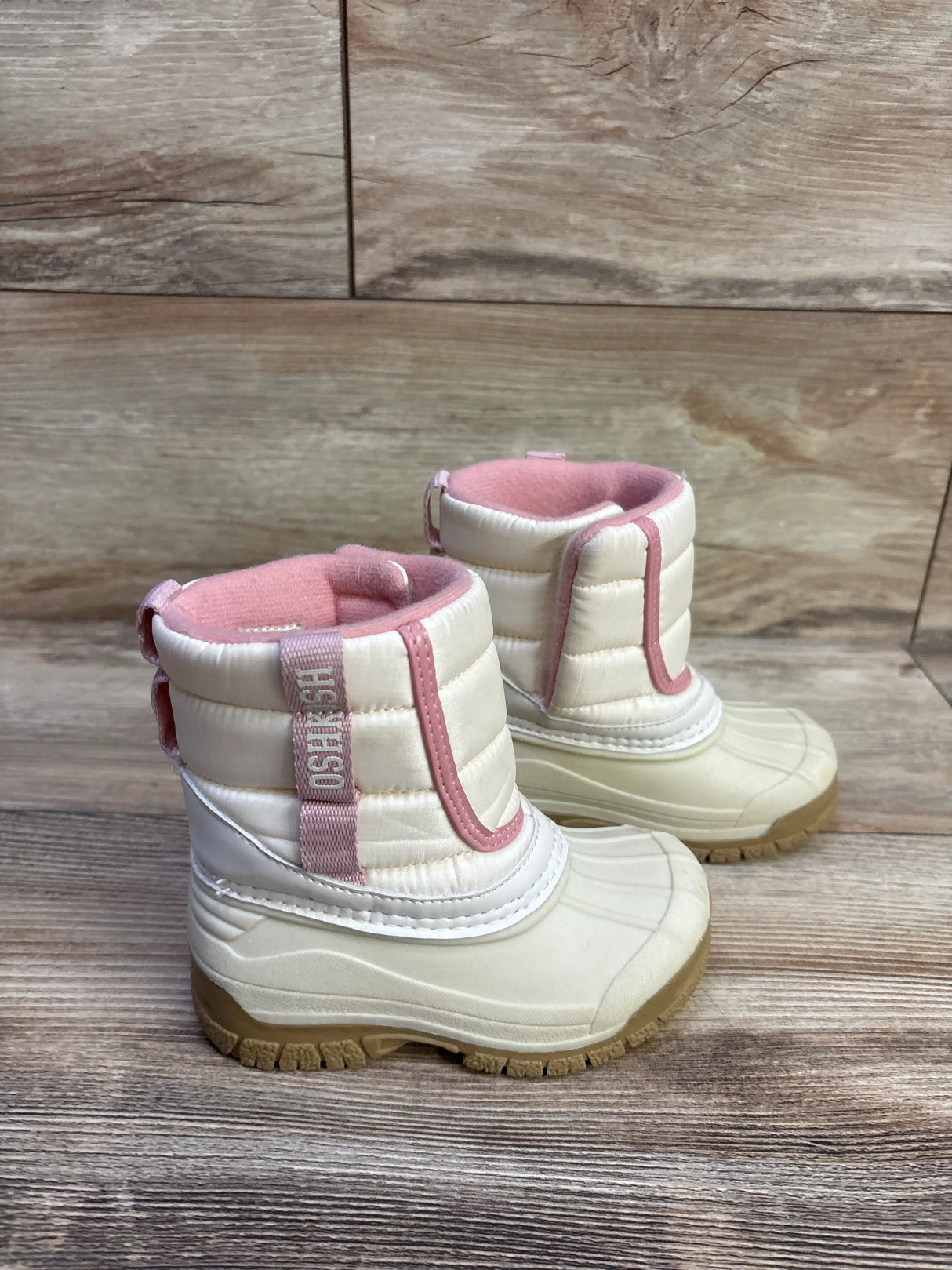 Side view of Oshkosh toddler ivory winter boots showing pink trim, fleece lining, and rubber soles on wood floor