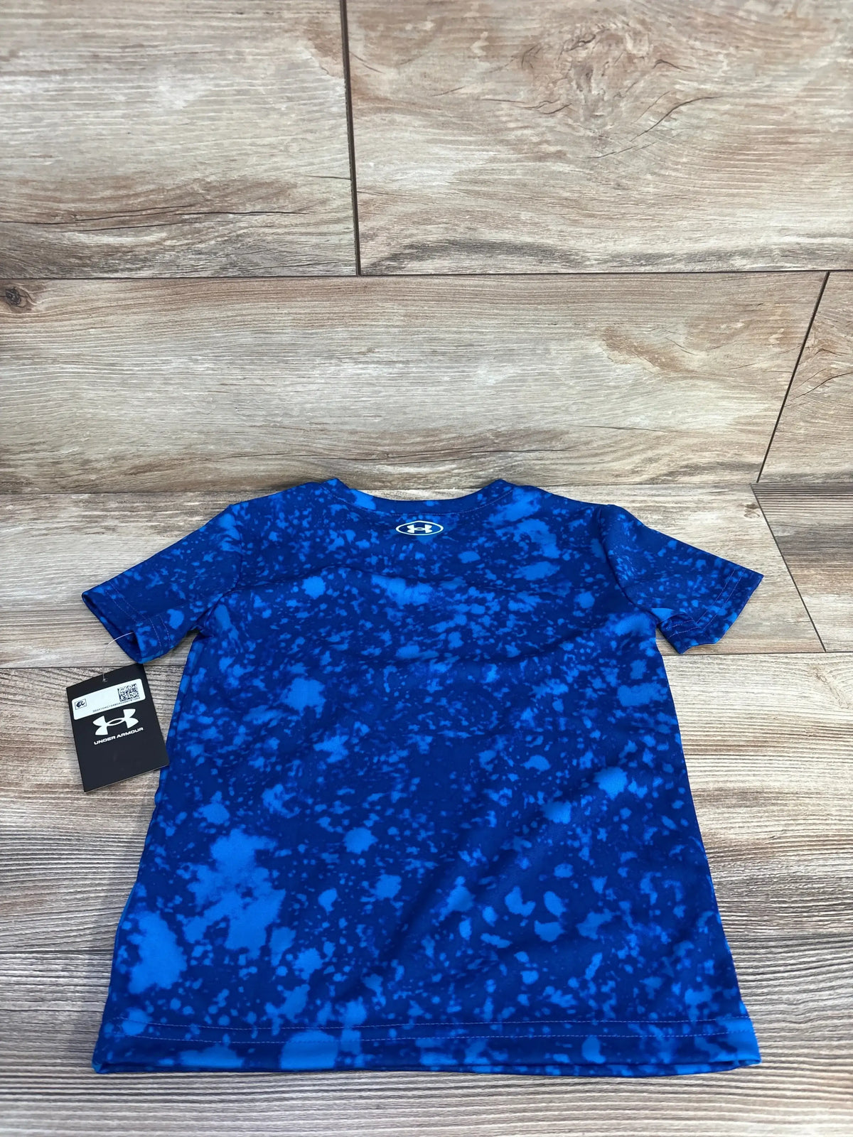 Flat lay of boys’ Under Armour short-sleeve active shirt 4T showcasing blue splatter print and back-neck logo with wood floor background