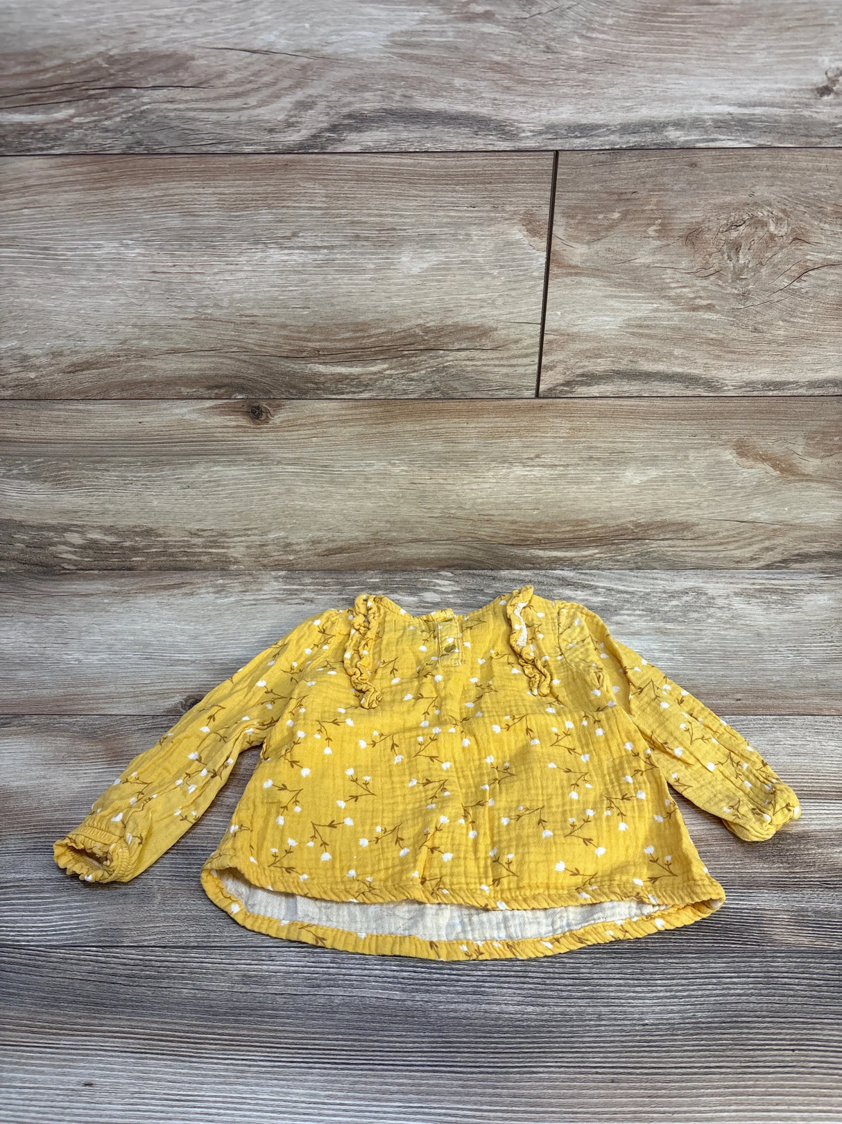 Yellow long sleeve baby shirt with floral pattern and ruffle details, laid on wood floor