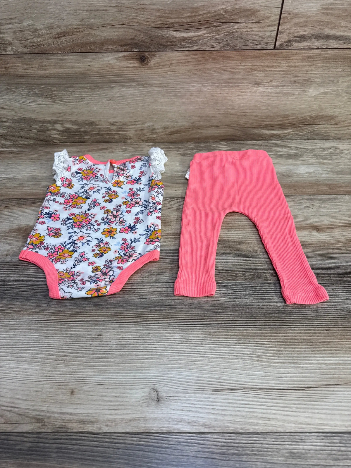 Flat lay of 2-piece baby set: white floral bodysuit with lace sleeves and coral ribbed leggings on wood background