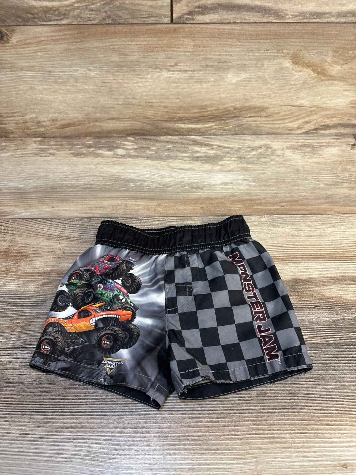 Front view of Monster Jam swim trunks, size 12m, showcasing black checkered print and monster truck graphics on wood