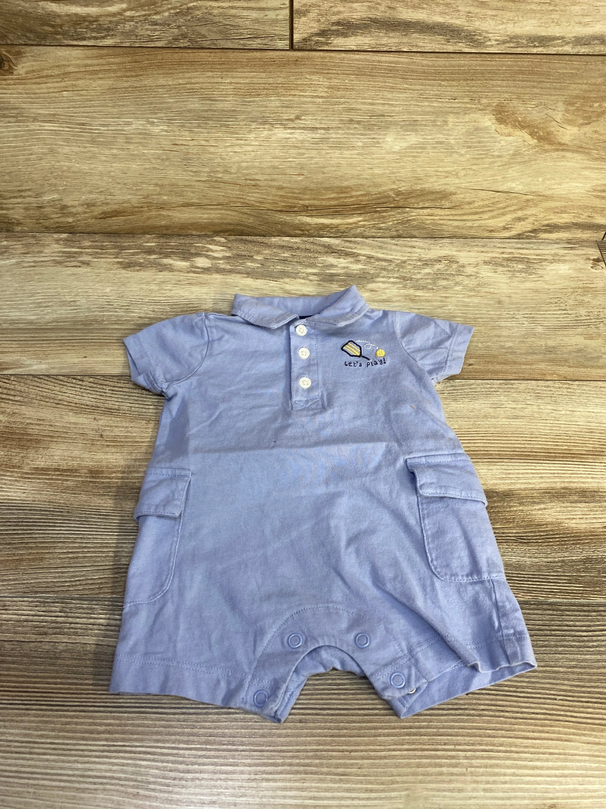 Top view of Carter's blue polo romper (3M) with short sleeves, button placket, side pockets and snap leg closures on wood
