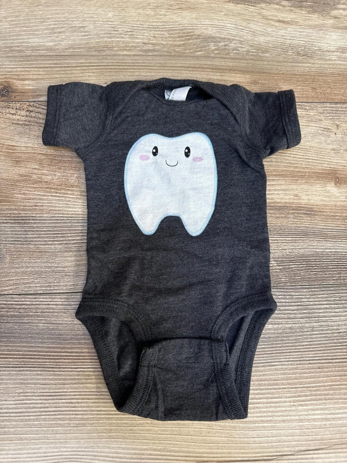 Flat lay of newborn charcoal baby bodysuit showcasing smiling tooth graphic on front with light wood background