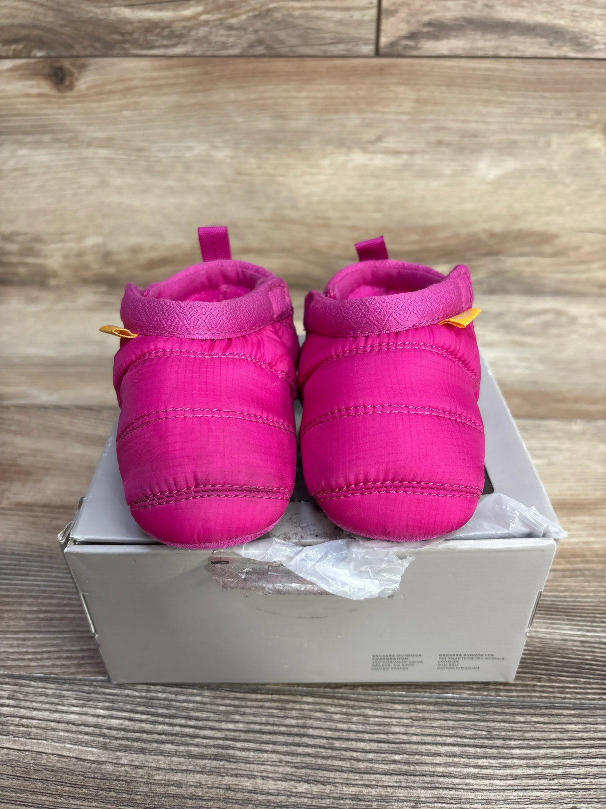 Front view of UGG Tasman LTA baby boots in bright pink with quilted insulated upper and pull tabs on a gray box and wood background