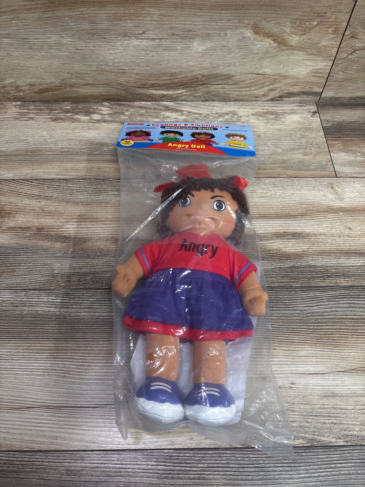 Front view of Angry washable doll in clear packaging on wood-grain surface, wearing red shirt labeled Angry