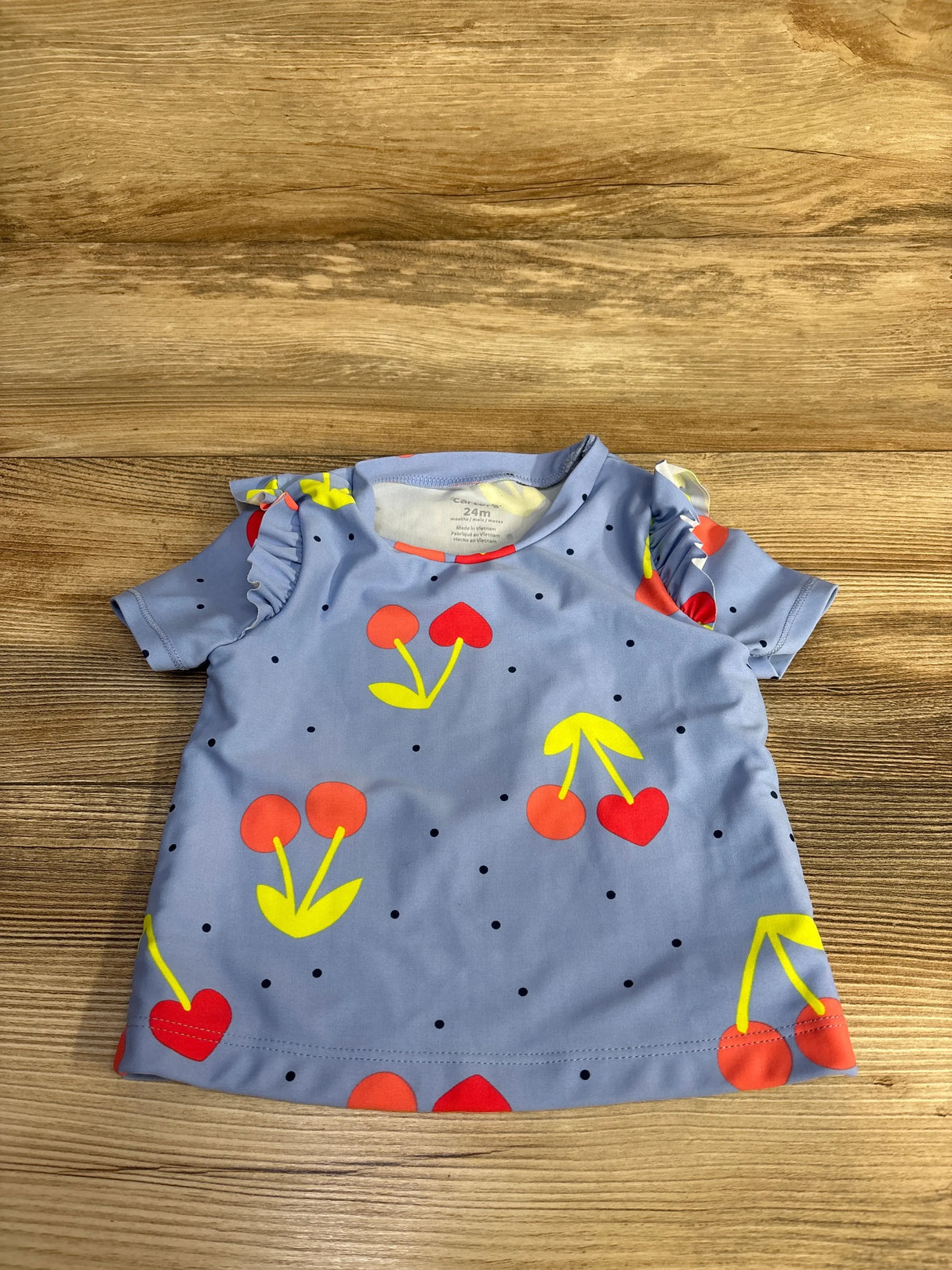 Front flat lay of Carter’s blue toddler rashguard (24M) showcasing red and yellow cherry print and ruffle shoulders on wood background